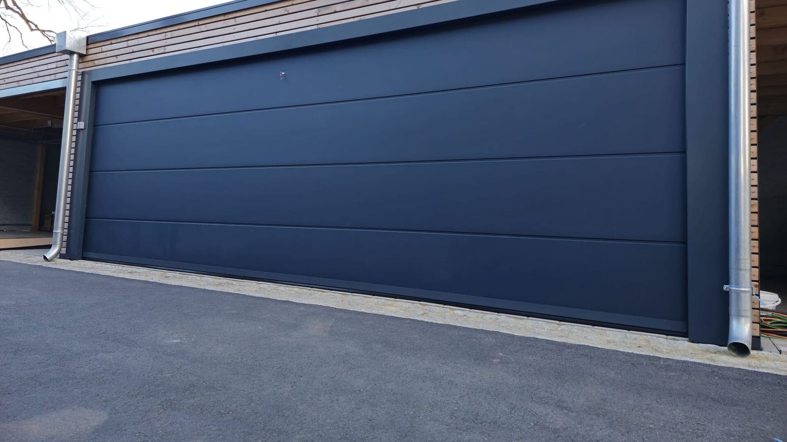 Très large porte de garage sectionnelle gris ardoise lisse sur un garage moderne bardé de bois