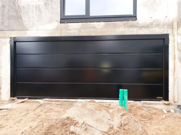 Breites schwarzes Sektionaltor mit breiten Sicken an einem Rohbau aus Sichtbeton