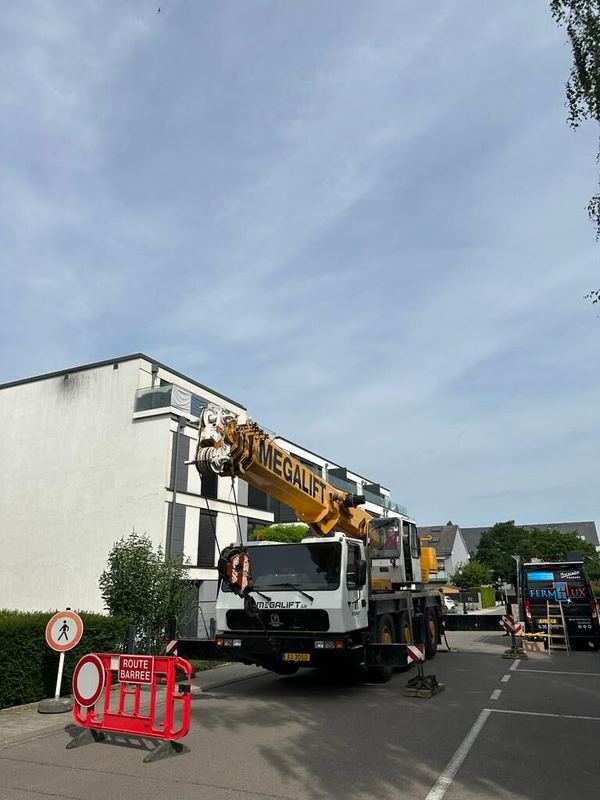Gelber Autokran vor einem modernen Wohnhaus, gesperrte Straße und Firmentransporter im Hintergrund