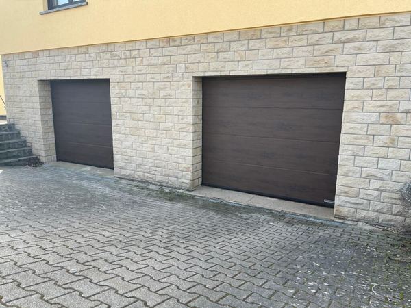 Zwei dunkelbraune Sektionaltore in einem hellen Natursteinsockel, gepflasterter Hof