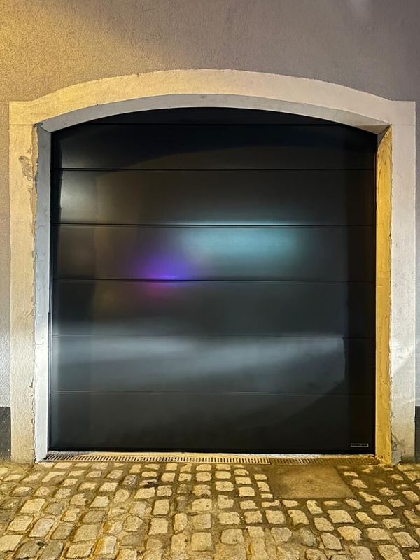 Glattes schwarzes Sektionaltor unter einem Rundbogen aus Stein, nachts über Kopfsteinpflaster