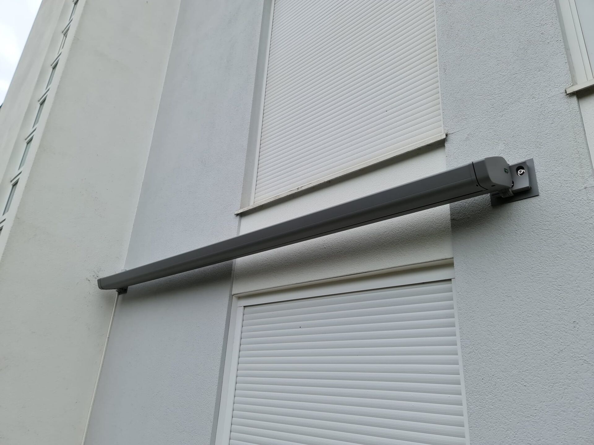 Compact grey awning cassette, closed, between two windows fitted with roller shutters