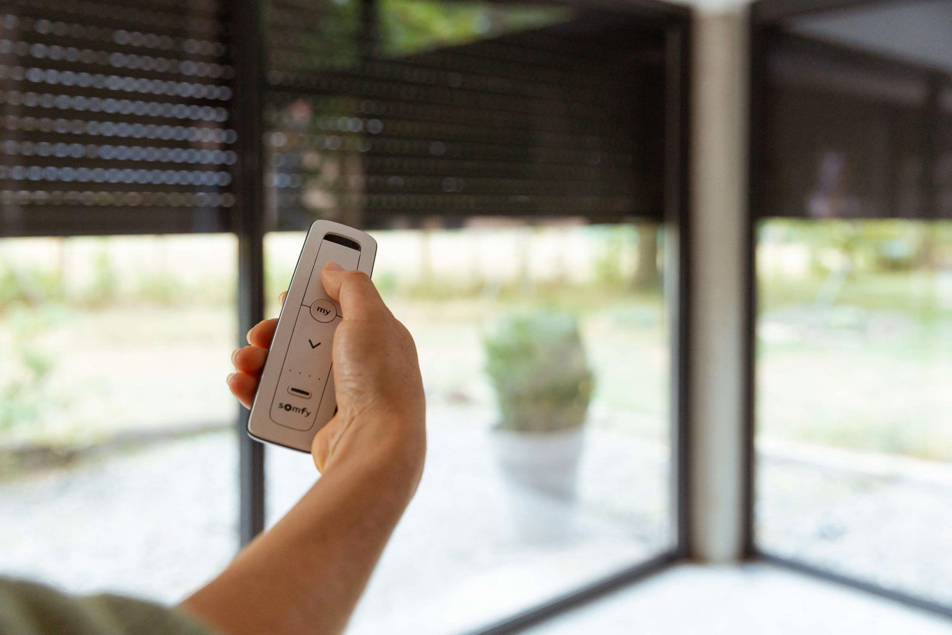 Hand mit einer Somfy-io-Fernbedienung, die dunkle Rollläden vor einer Glasfront herunterfährt