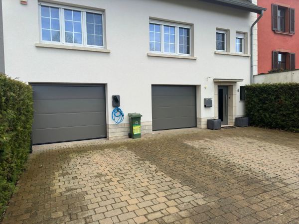 Zwei graue Sektionaltore an einem weißen Wohnhaus, Wallbox und gepflasterter Hof
