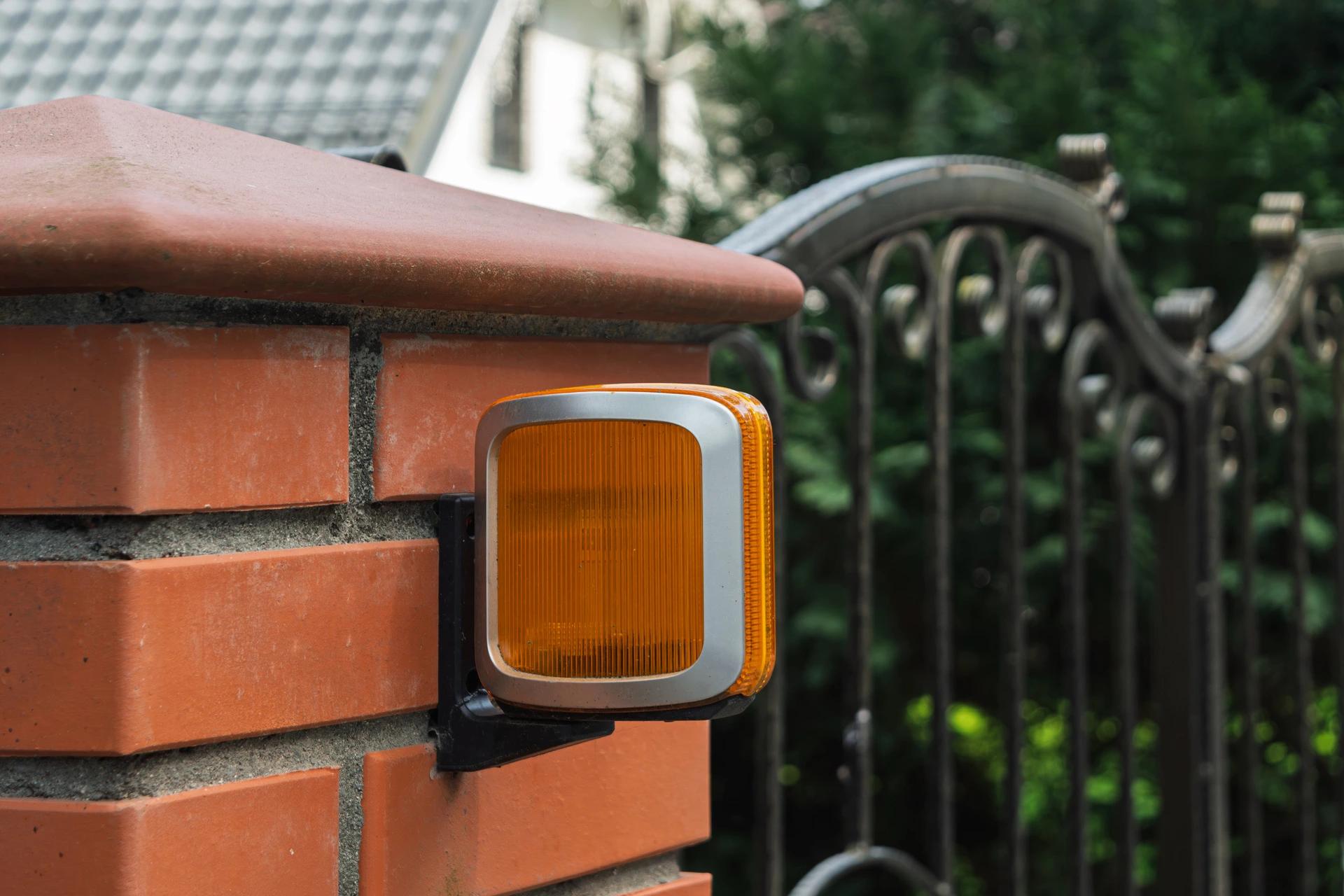 Orange gate motor warning light mounted on a brick pillar beside a wrought-iron gate