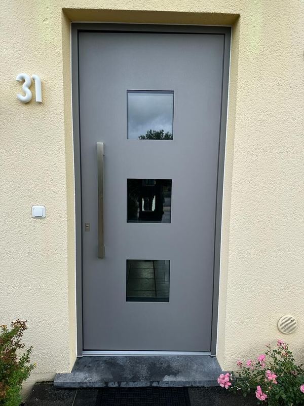 Hellgraue Aluminium-Haustür mit drei versetzten quadratischen Glasfeldern und langem Edelstahlgriff, cremefarbener Putz
