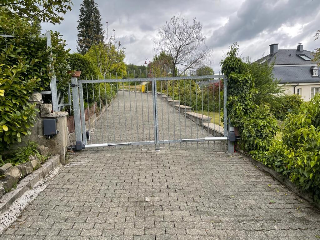 Zweiflügeliges Drehtor aus verzinktem Stahl mit Gelenkarmantrieb an einer gepflasterten Zufahrt