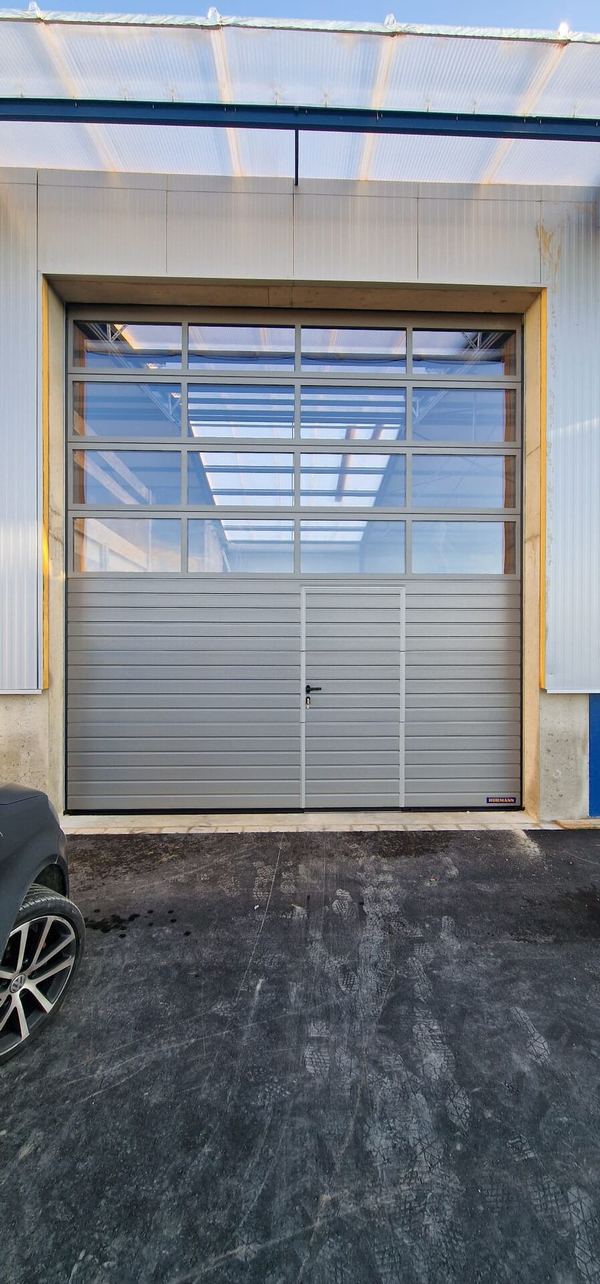 Graues Industrie-Sektionaltor mit 4 verglasten Reihen und Schlupftür unter einem Lichtdach