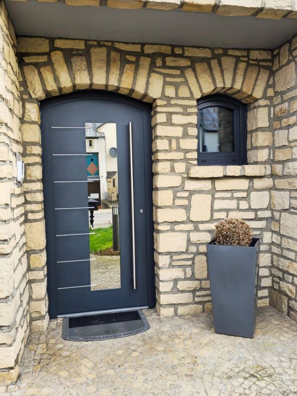 Anthrazitfarbene Haustür mit Rundbogen, senkrechter Verglasung, Edelstahl-Applikationen und Stangengriff, Natursteinfassade
