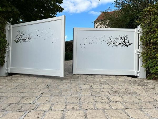 Weißes zweiflügeliges Drehtor, halb geöffnet, mit ausgeschnittenem Ast- und Blattmotiv, Pflasterzufahrt
