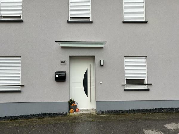 Weiße Haustür mit Halbmond-Glas unter Glasvordach, graue Fassade mit weißen Rollläden
