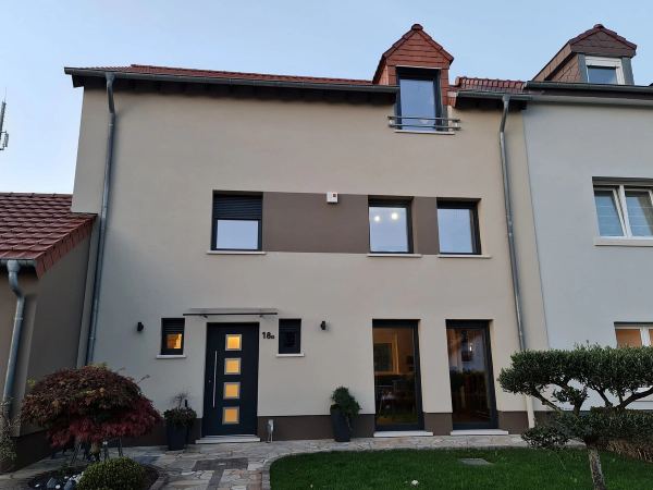 Beige-taupefarbene Fassade in der Dämmerung, anthrazitfarbene Fenster, zwei bodentiefe Fenster und Haustür
