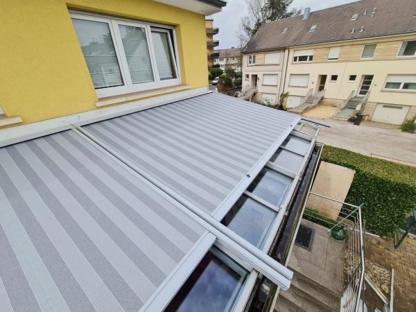 Grau-weiß gestreifte Wintergartenmarkise auf Glasdach, gelbes Haus von oben
