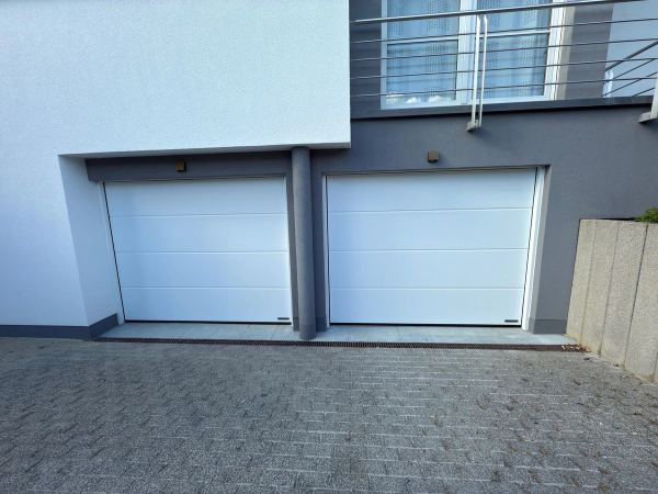 Zwei weiße Sektionaltore unter einer Terrasse mit Edelstahlgeländer, weiß-graue Fassade