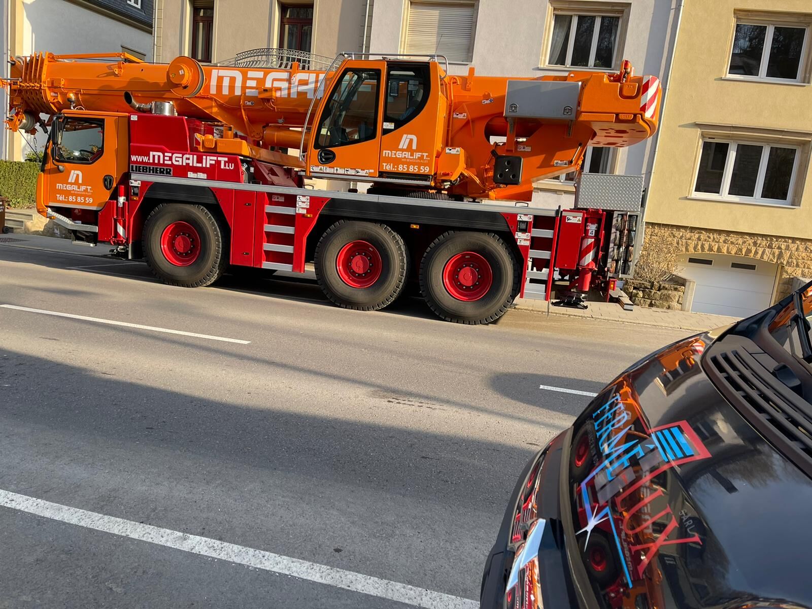 Orangefarbener Autokran in einer Wohnstraße für eine Baustellenlieferung, Motorhaube eines Transporters im Vordergrund