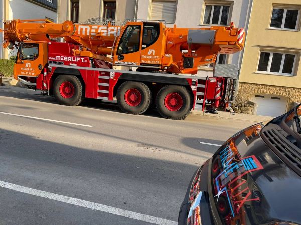 Orangefarbener Autokran in einer Wohnstraße für eine Baustellenlieferung, Motorhaube eines Transporters im Vordergrund