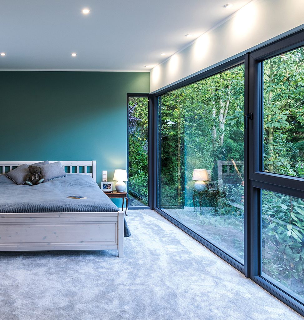 Schlafzimmer mit großen anthrazitgrauen Kunststoff-Eckfenstern mit Blick in einen begrünten Garten