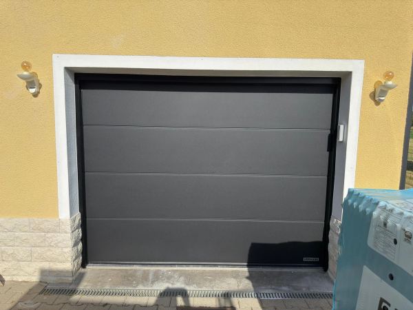 Anthrazitfarbenes Sektionaltor zwischen zwei Wandleuchten an gelber Fassade mit weißer Umrandung