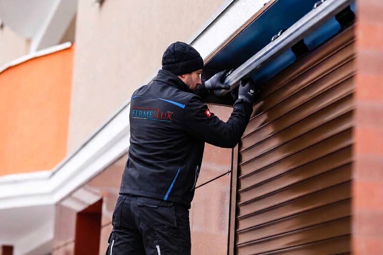 Fermé-Lux technician working on the motorised shaft of a brown electric roller shutter on a façade