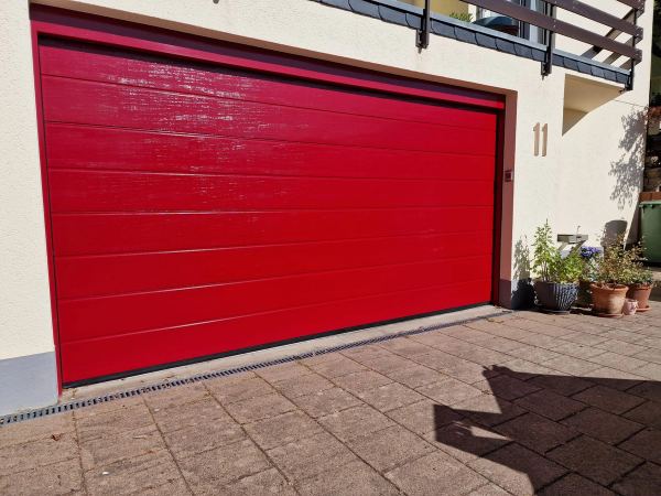 Leuchtend rotes Doppel-Sektionaltor mit Sicken, cremefarbene Putzfassade und gepflasterte Einfahrt