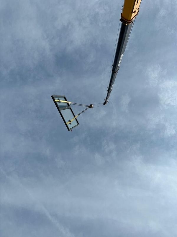 Großes Glaselement mit dunklem Rahmen am Ausleger eines Krans vor bewölktem Himmel
