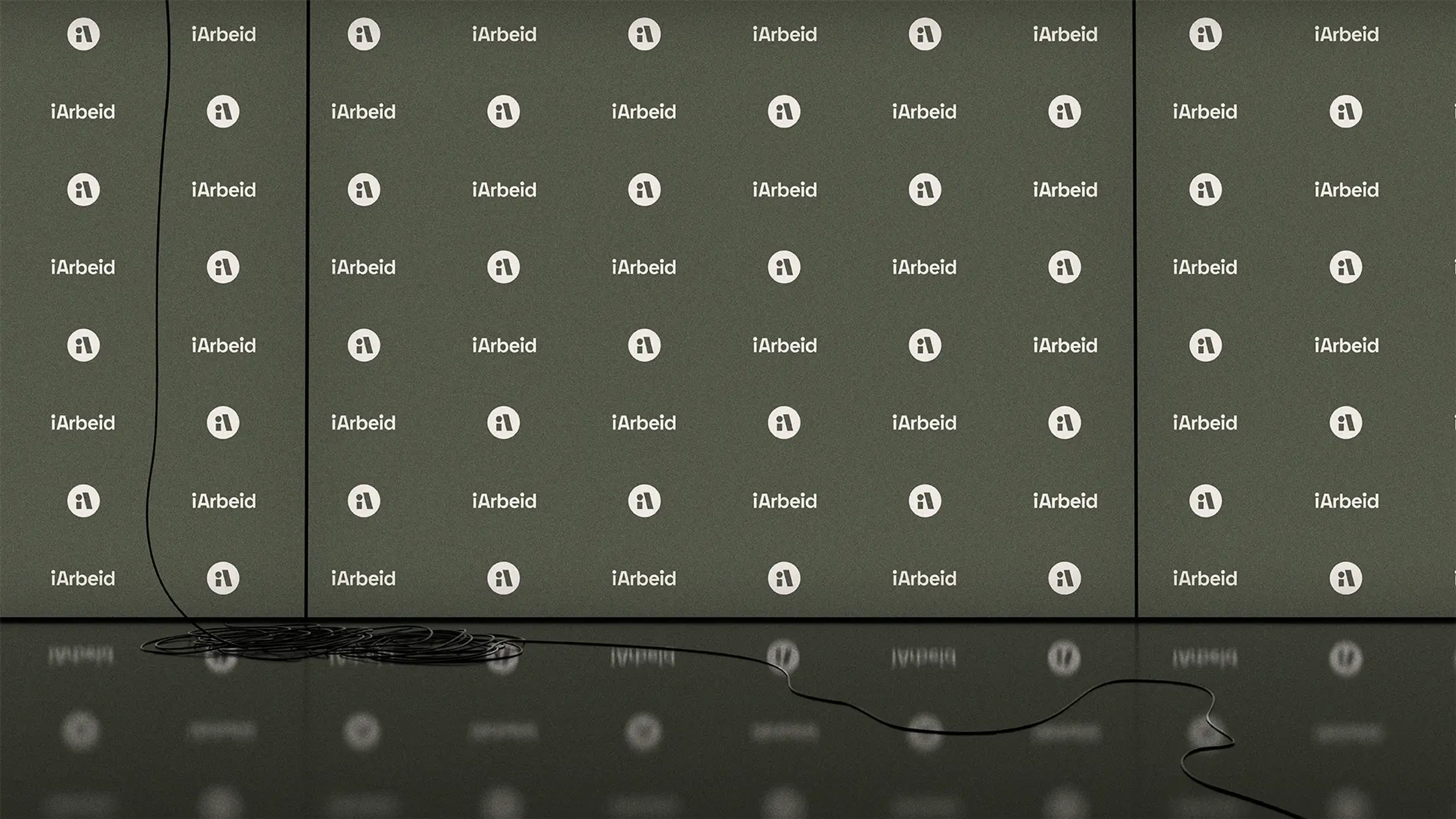 En mørkegrønn pressevegg med et repeterende mønster av logoen og navnet "iArbeid" i hvitt. Veggen består av tre paneler, og foran den er det et mørkt, speilende gulv. En tynn, svart ledning henger ned fra venstre side og ligger i en kveil på gulvet, før den strekker seg utover mot høyre side av bildet.