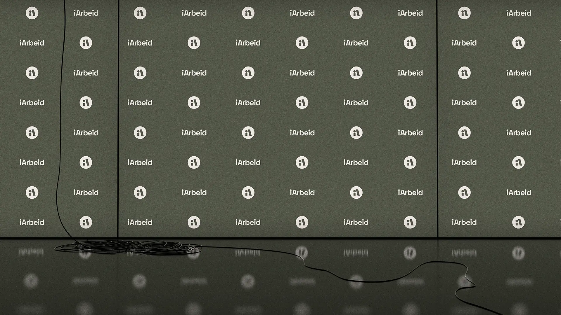 En mørkegrønn pressevegg med et repeterende mønster av logoen og navnet "iArbeid" i hvitt. Veggen består av tre paneler, og foran den er det et mørkt, speilende gulv. En tynn, svart ledning henger ned fra venstre side og ligger i en kveil på gulvet, før den strekker seg utover mot høyre side av bildet.
