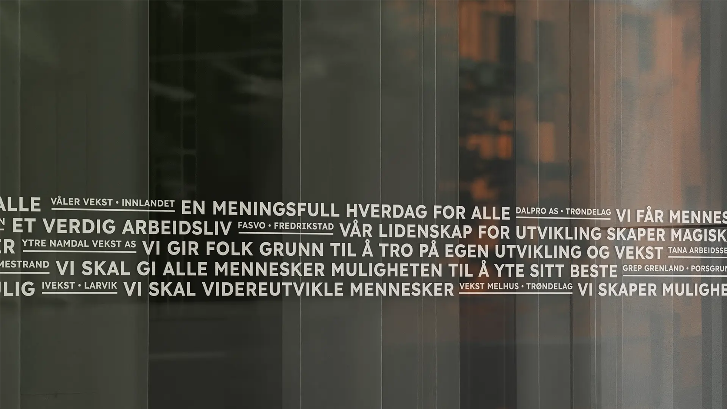 Nærbilde av en glassvegg med hvit tekst som presenterer ulike slagord og firmanavn. Teksten er skrevet i blokkbokstaver og inkluderer setninger som «En meningsfull hverdag for alle», «Et verdig arbeidsliv» og «Vi skal gi alle mennesker muligheten til å yte sitt beste». Mellom slagordene står navn på ulike vekstbedrifter og steder, som «Våler Vekst • Innlandet» og «Vekst Melhus • Trøndelag». Bakgrunnen er mørk og reflektorerer et interiør med varme toner.