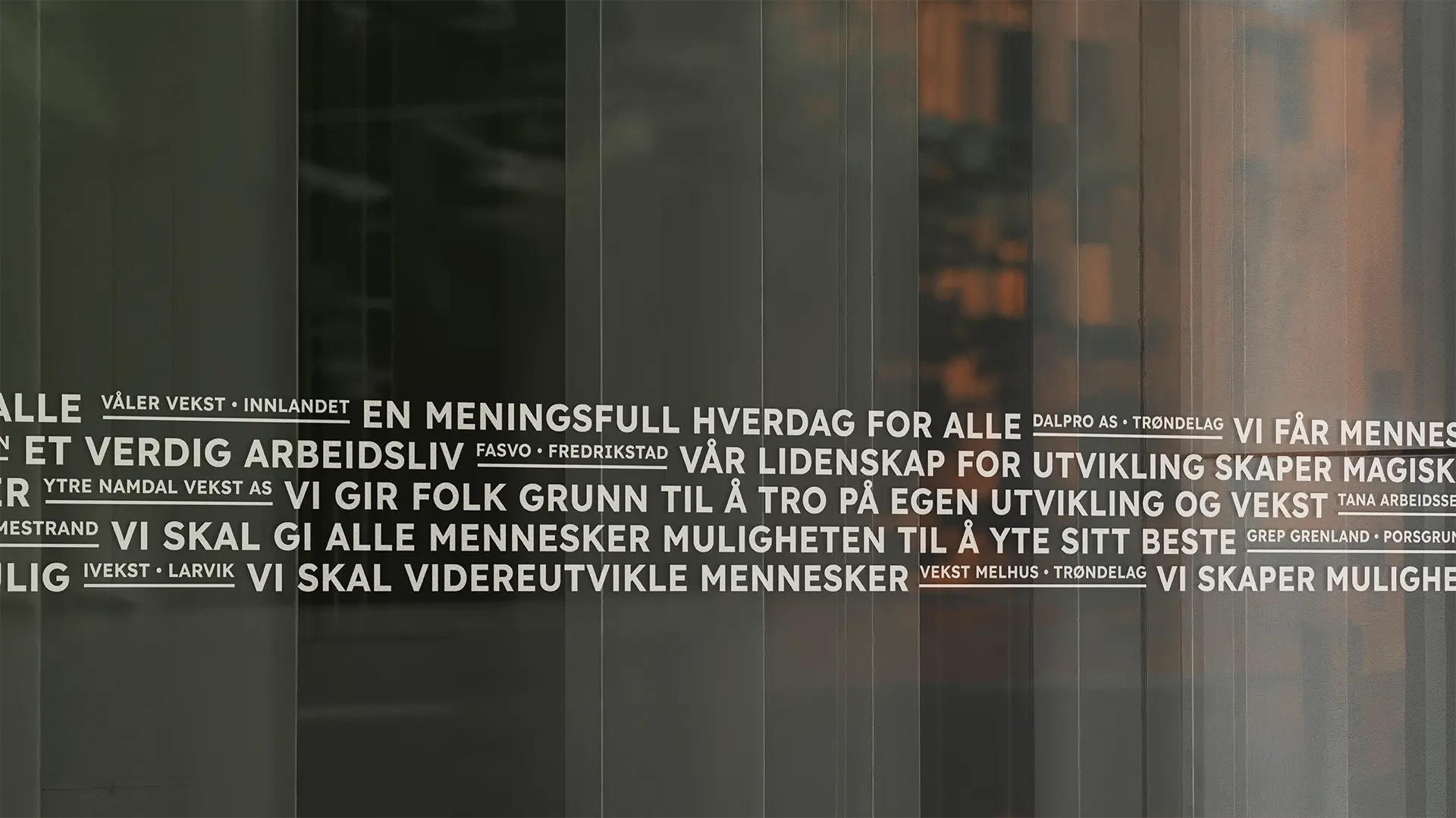 Nærbilde av en glassvegg med hvit tekst som presenterer ulike slagord og firmanavn. Teksten er skrevet i blokkbokstaver og inkluderer setninger som «En meningsfull hverdag for alle», «Et verdig arbeidsliv» og «Vi skal gi alle mennesker muligheten til å yte sitt beste». Mellom slagordene står navn på ulike vekstbedrifter og steder, som «Våler Vekst • Innlandet» og «Vekst Melhus • Trøndelag». Bakgrunnen er mørk og reflektorerer et interiør med varme toner.