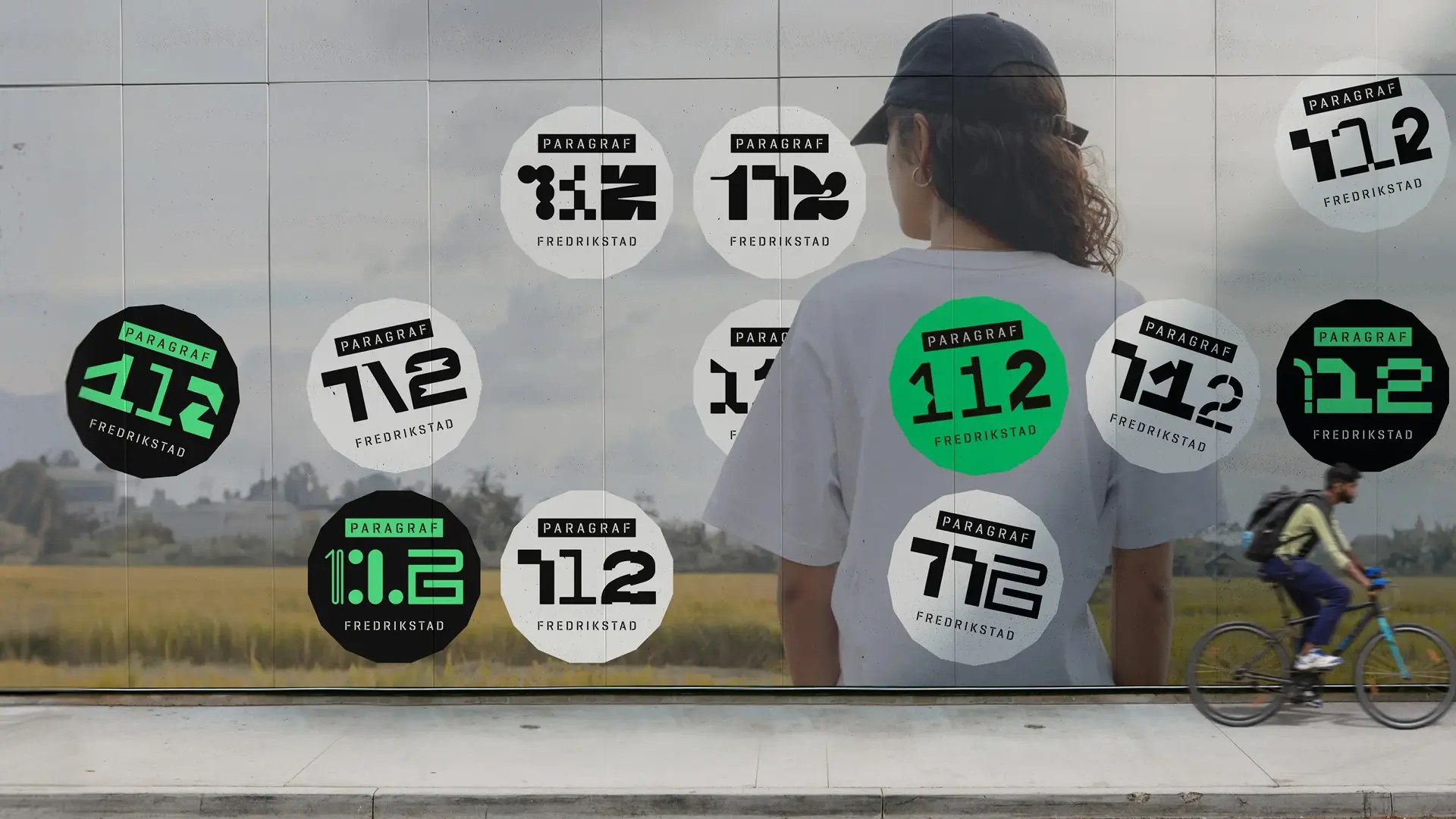 En utendørs vegg dekket med ulike varianter av Paragraf 112-logoen i sirkler, med en person og en syklist i forgrunnen.