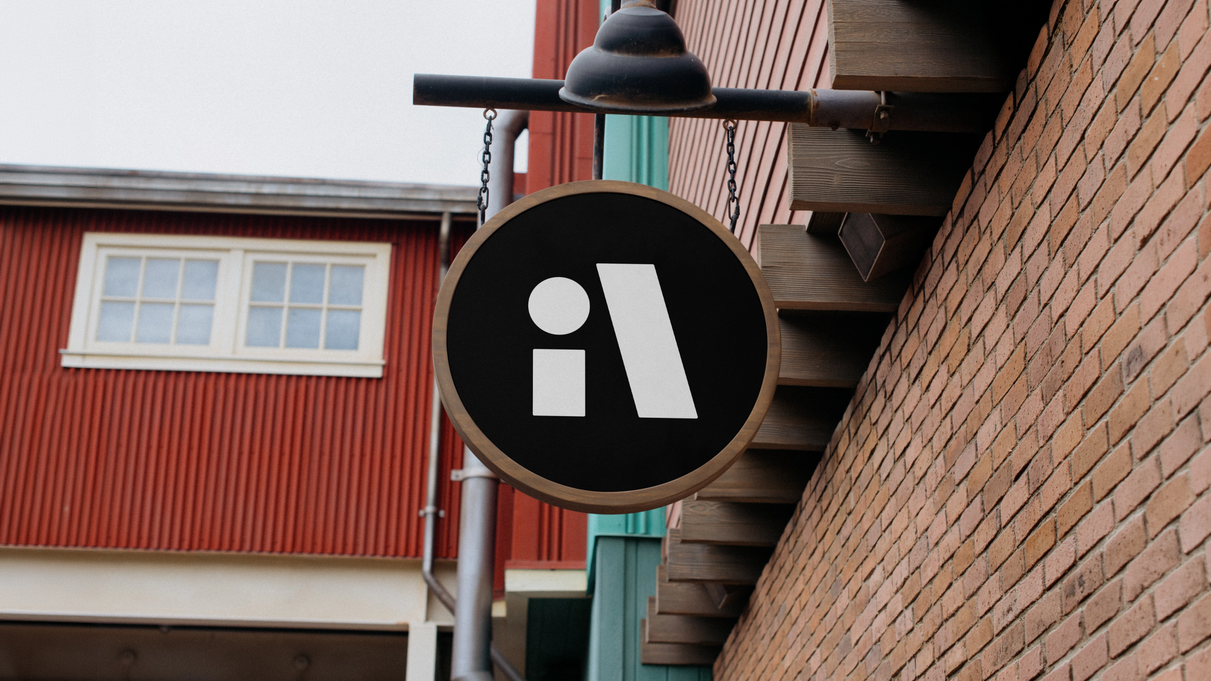 Et rundt, svart skilt med en hvit logo hengende fra en husvegg av mur og rød panel. Logoen består av en stilisert, liten bokstav «i» ved siden av en tykk, skråstilt blokk som danner bokstaven «A».