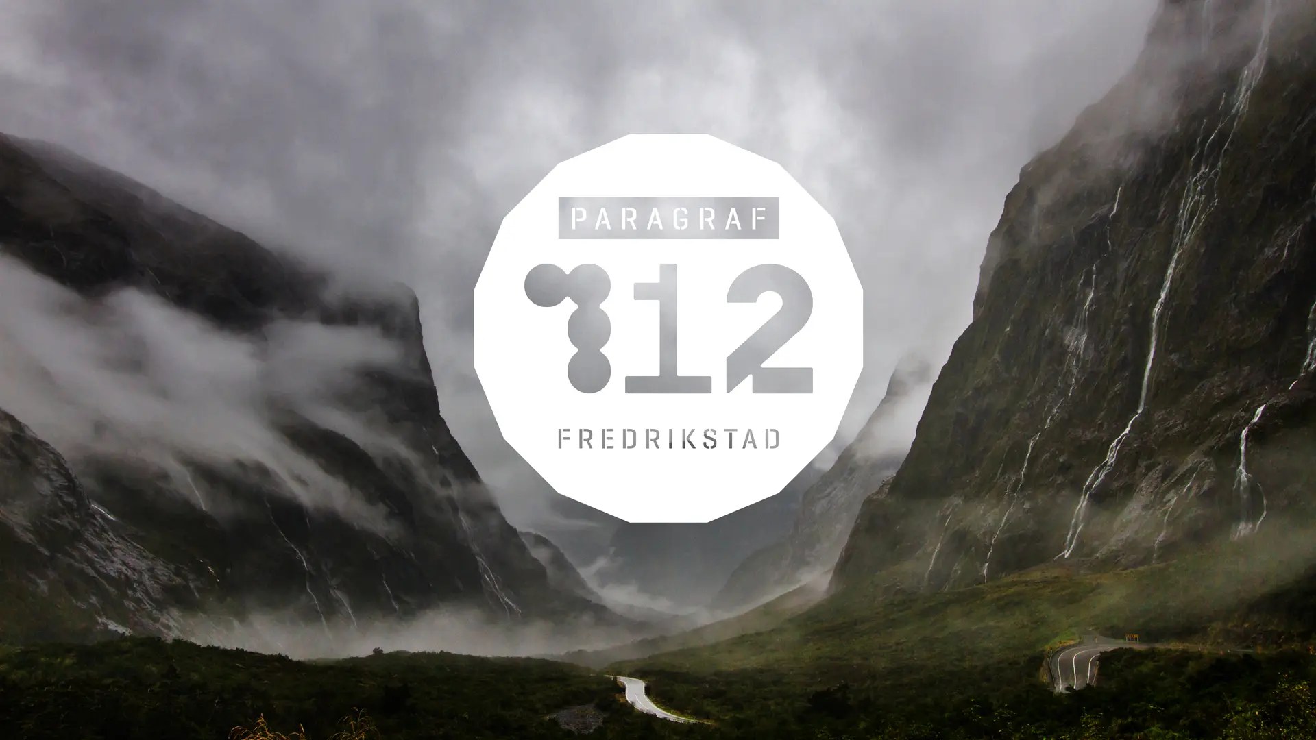 En stor, hvit sirkulær logo med teksten «PARAGRAF 112 FREDRIKSTAD» er plassert over et dramatisk bilde av en dyp dal. Tåke og lave skyer driver mellom bratte fjellsider med små fosser, og en smal vei snirkler seg gjennom bunnen av dalen.