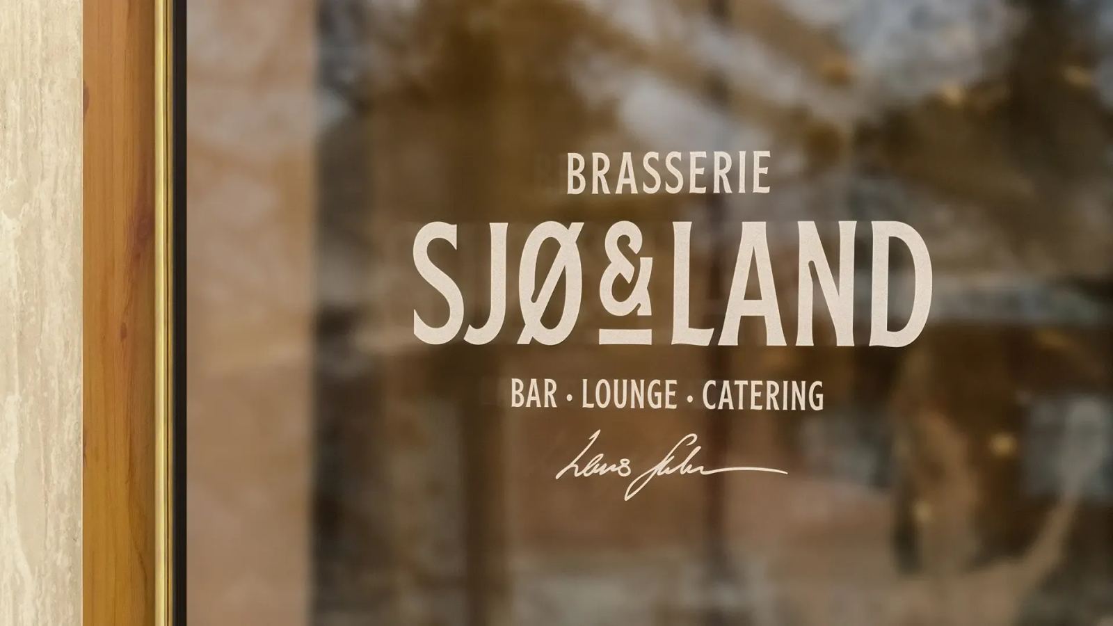 Logoen til Brasserie Sjø & Land i lys folie på et vindu, med «Bar · Lounge · Catering».