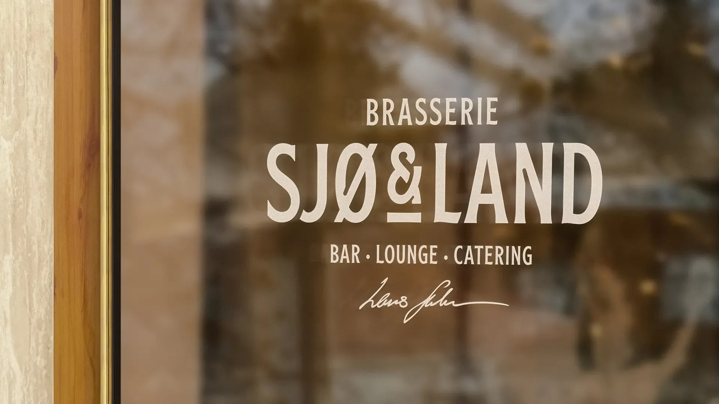 Logoen til Brasserie Sjø & Land i lys folie på et vindu, med «Bar · Lounge · Catering» og en håndskrevet signatur under.