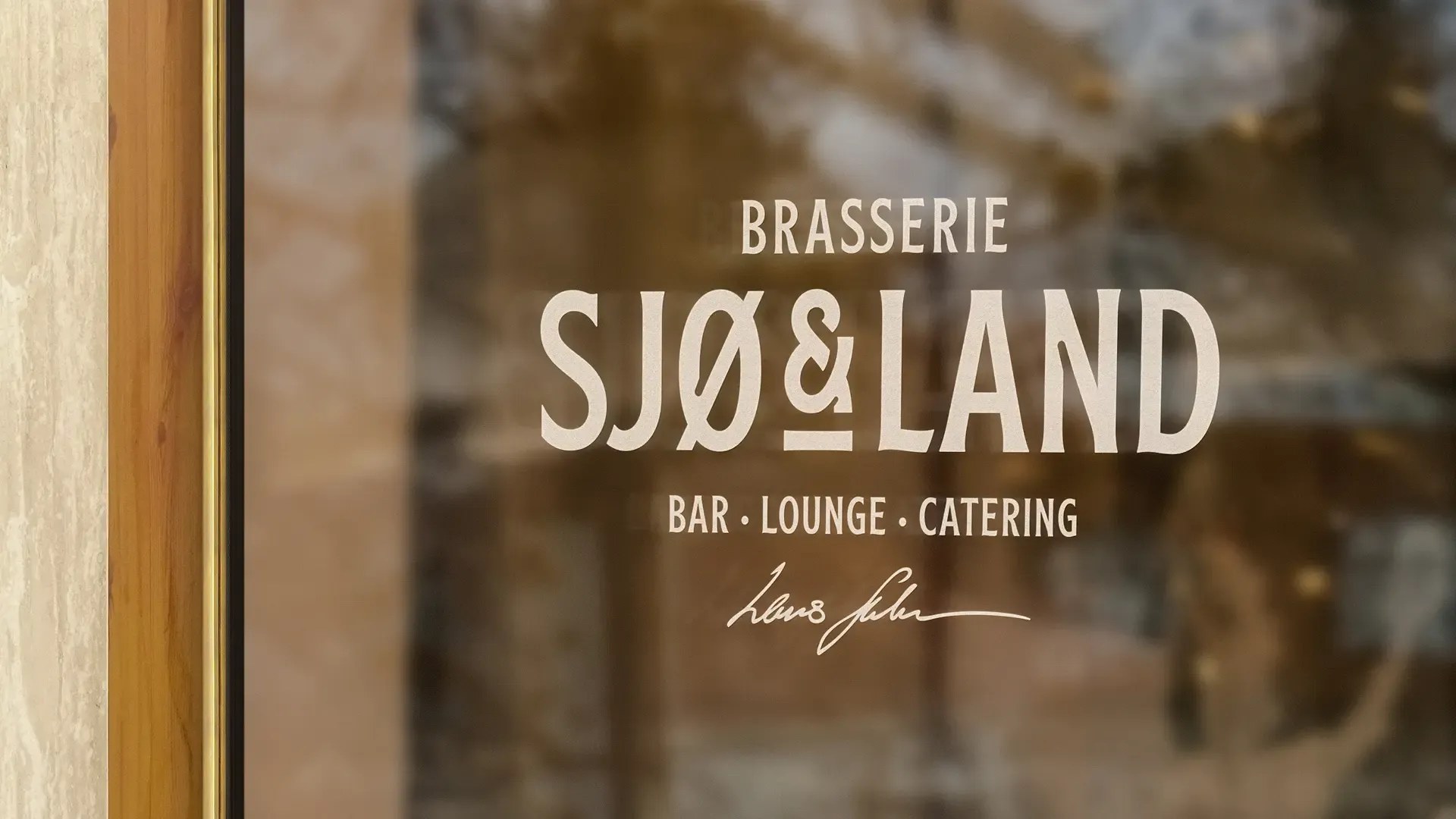 Logoen til Brasserie Sjø & Land i lys folie på et vindu, med «Bar · Lounge · Catering» og en håndskrevet signatur under.