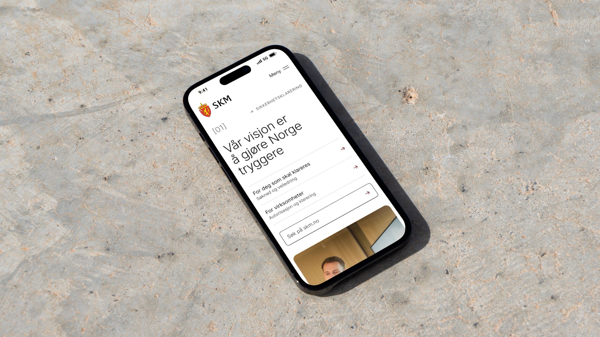 En smarttelefon som viser nettsiden til SKM (Sivil klareringsmyndighet) med visjonen «Vår visjon er å gjøre Norge tryggere», plassert på en grå betongbakgrunn.