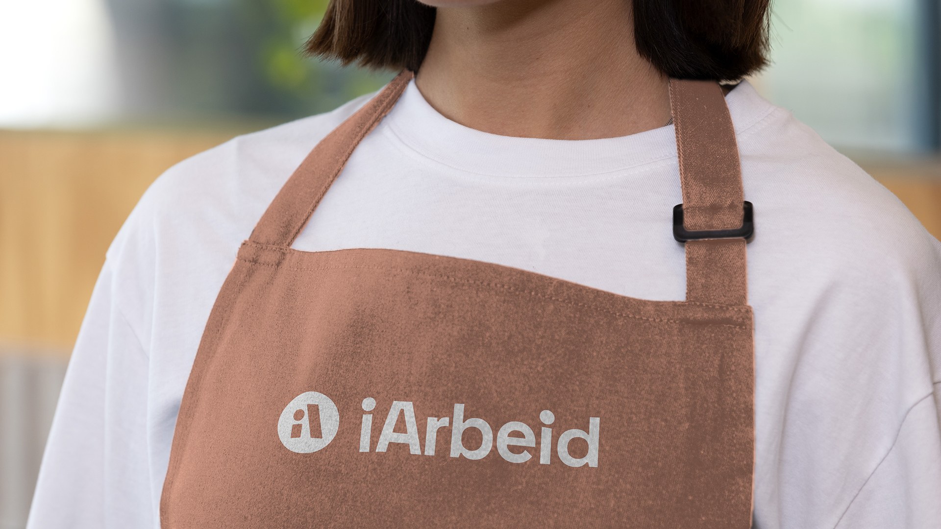 Nærbilde av en person som bærer et brunt forkle med iArbeid-logoen trykket i hvitt på brystet.