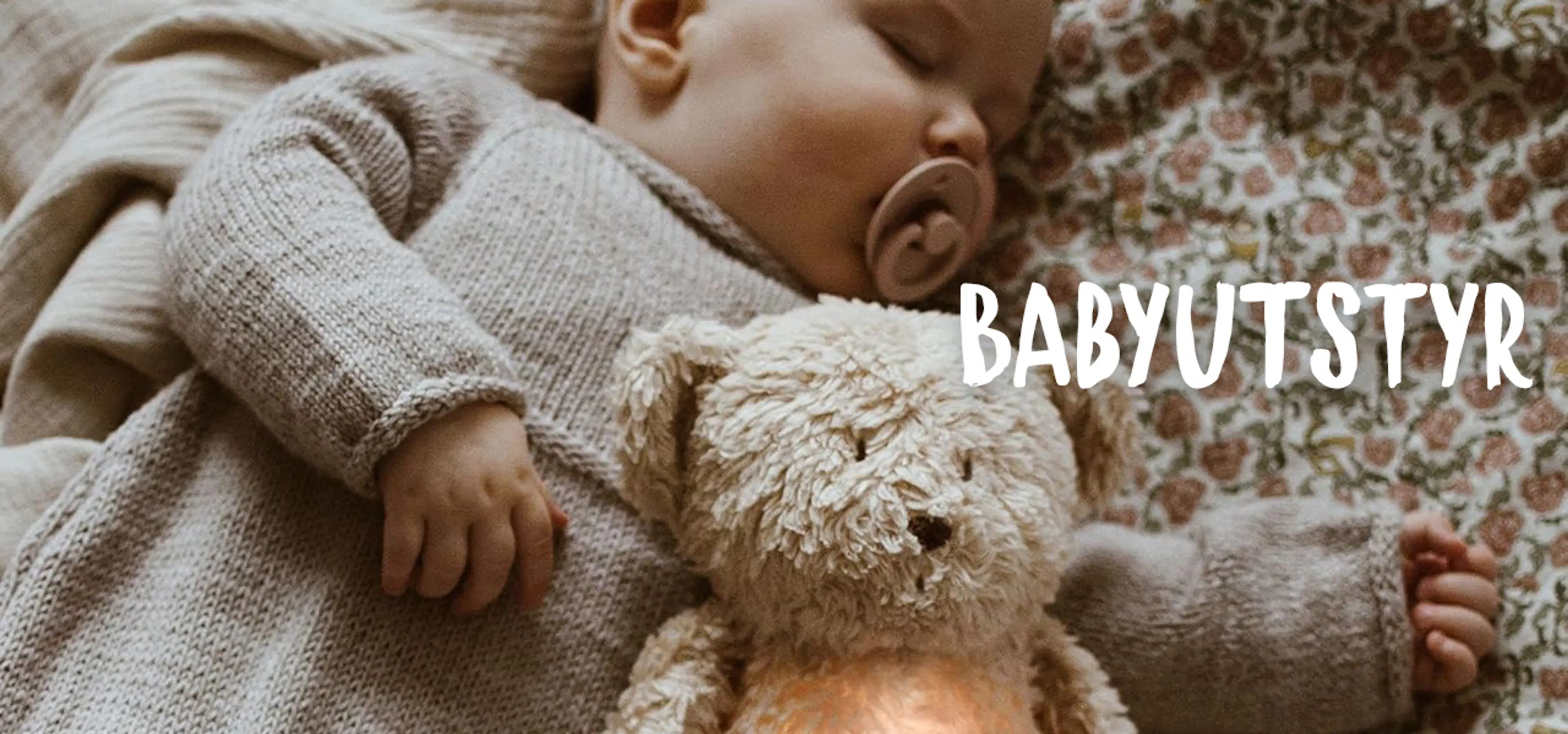 Babyutstyr. Fotografi av en sovende baby med en Moonie bamse i hendene.