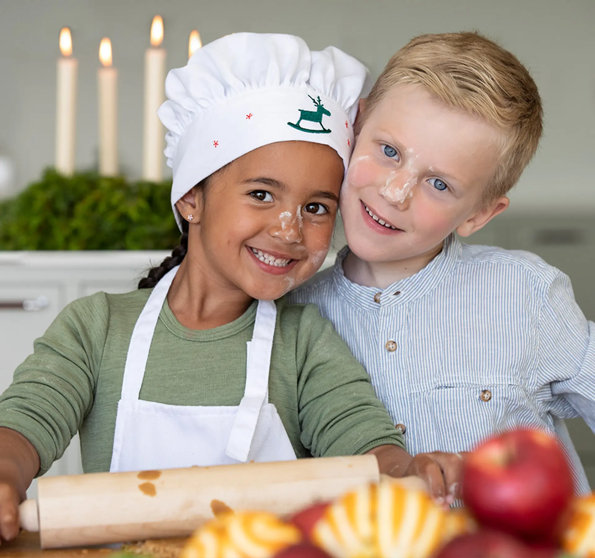 Fotografi av to smilende barn med kokkelue, kjevle og bakst i ansiktet. Trykk videre for å se mer til julebaksten.