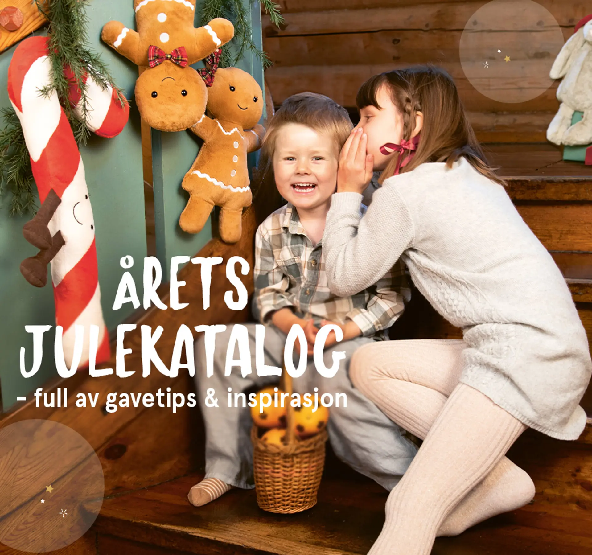 "Årets julekatalog er her - full av gavetips og inspirasjon!" Julekatalogen er avbildet over forsidebildet av to hviskende barn sittende i trapp med Jellycat Amuseables i bakgrunnen.