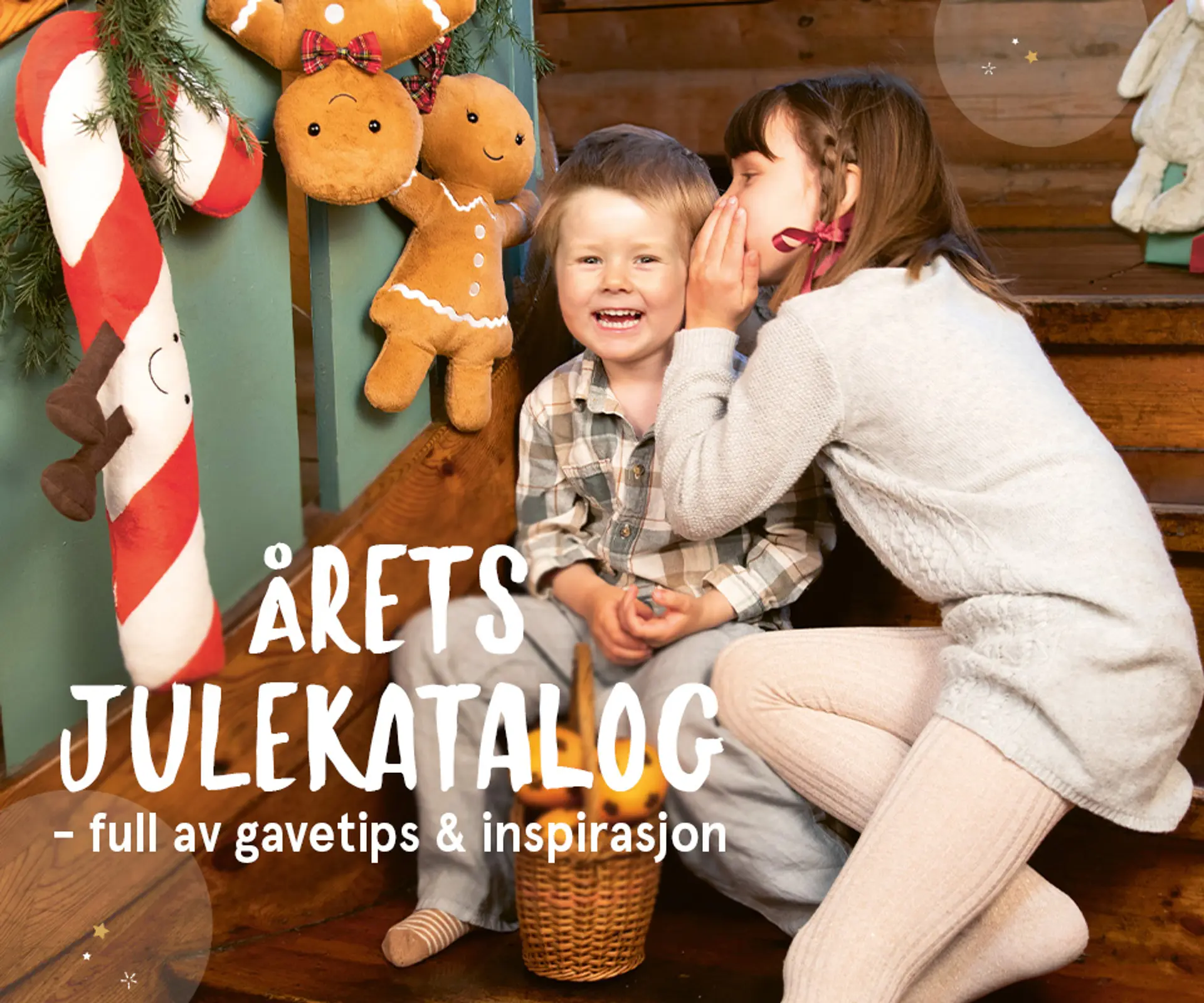 "Årets julekatalog er her - full av gavetips og inspirasjon!" Julekatalogen er avbildet over forsidebildet av to hviskende barn sittende i trapp med Jellycat Amuseables i bakgrunnen.