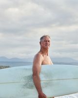 An old male surfer looking at you. He is on the beach. It is an overcast day. He has his surfboard in his hand.