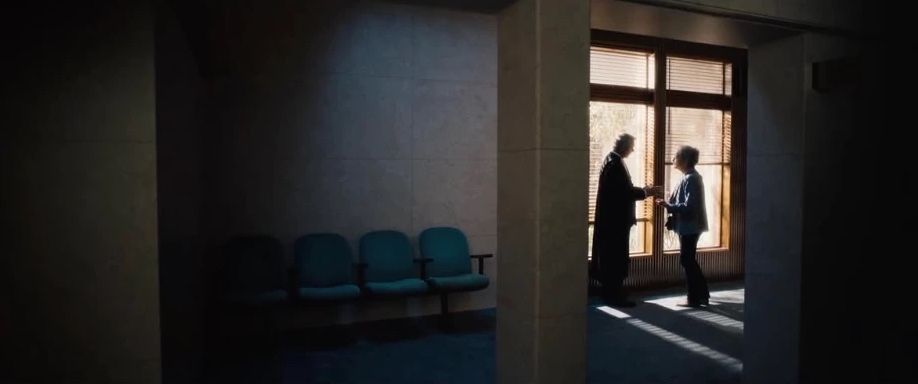 Two figures talk against a sunlit window at the end of a dark corridor lined with empty chairs.