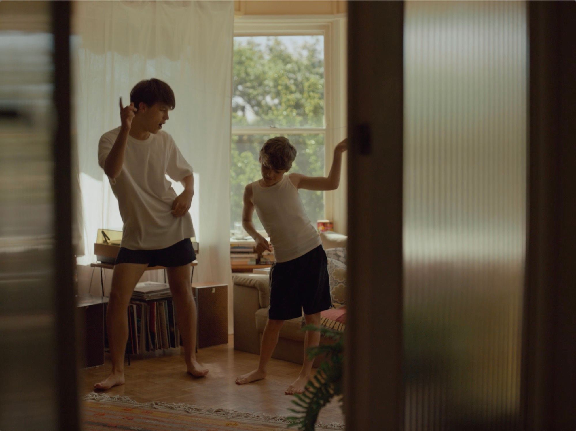 Two boys dance in a sunlit bedroom, seen through a doorway.