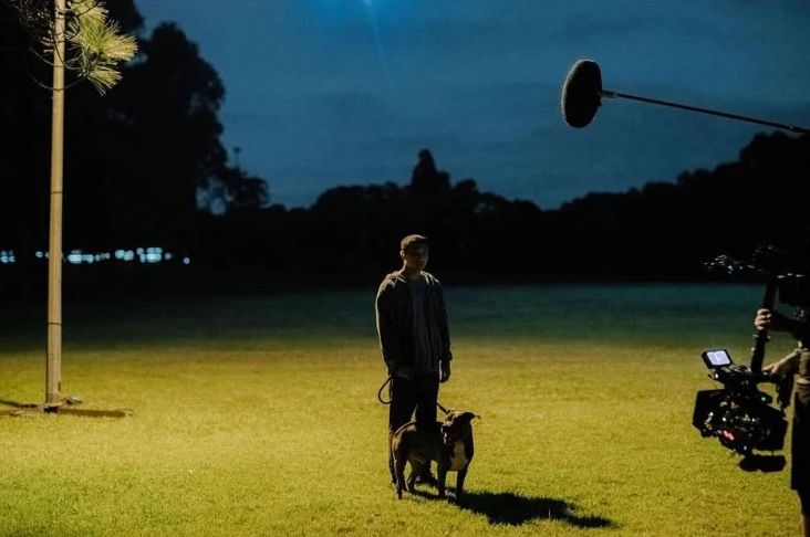 Behind the scenes: an actor with a dog stands on a lit oval at dusk as the camera and boom close in.
