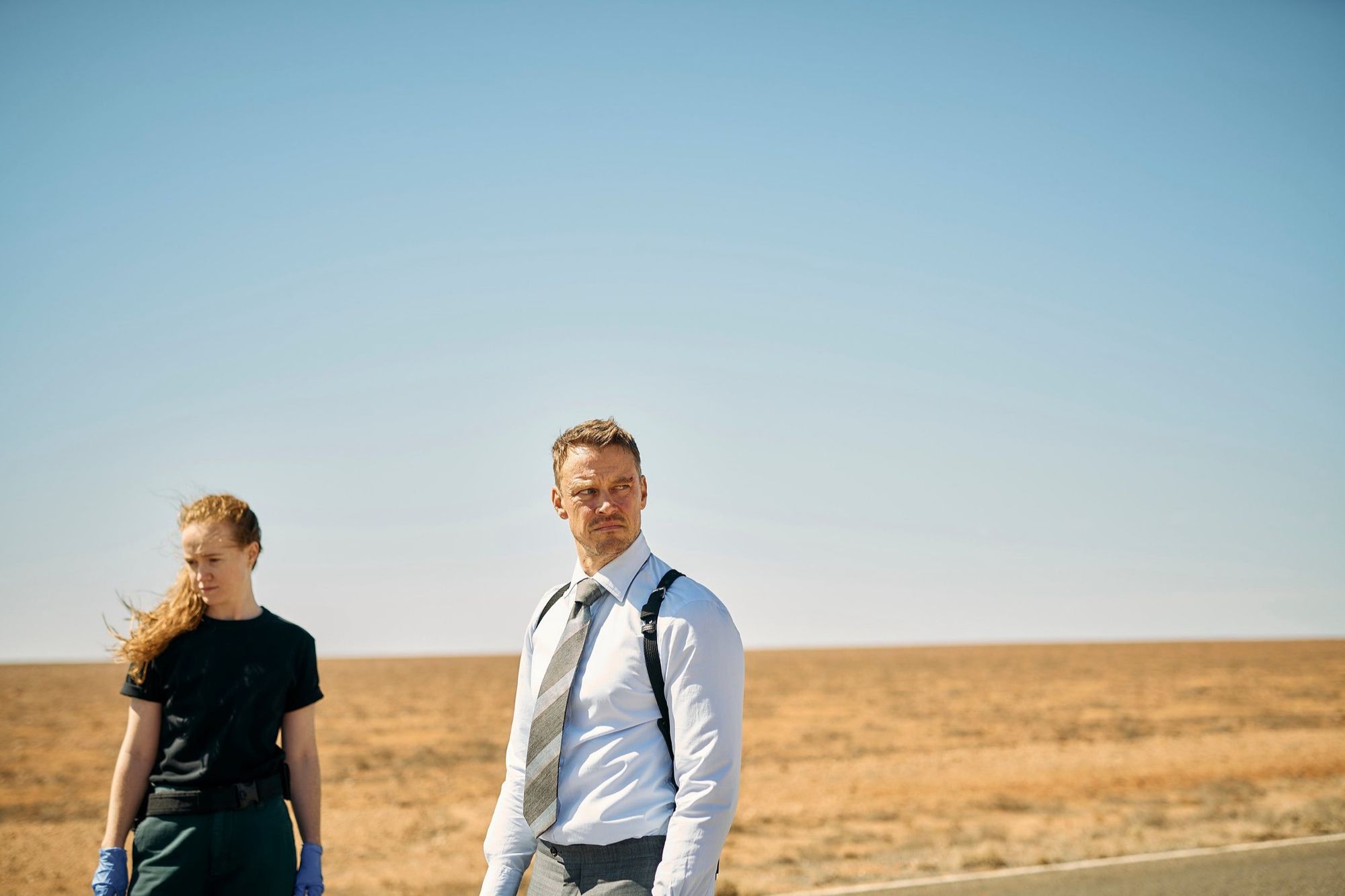 Two figures stand at the edge of a vast flat desert under a pale sky.