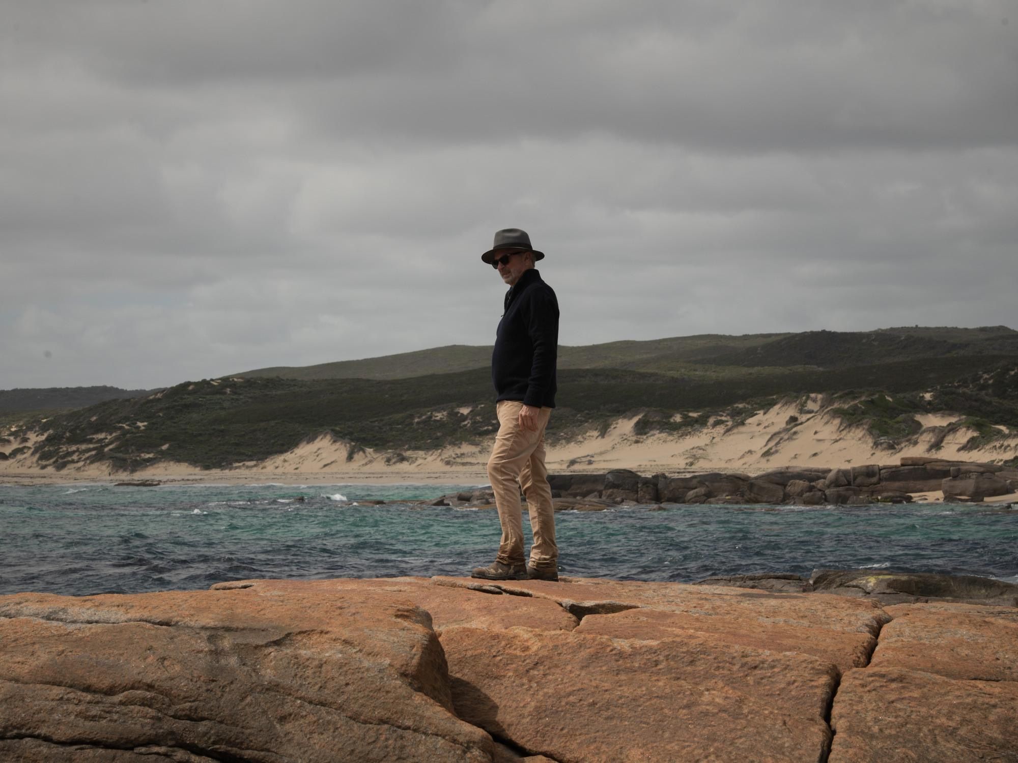 Sam Neill stands on orange coastal rocks under a grey sky.