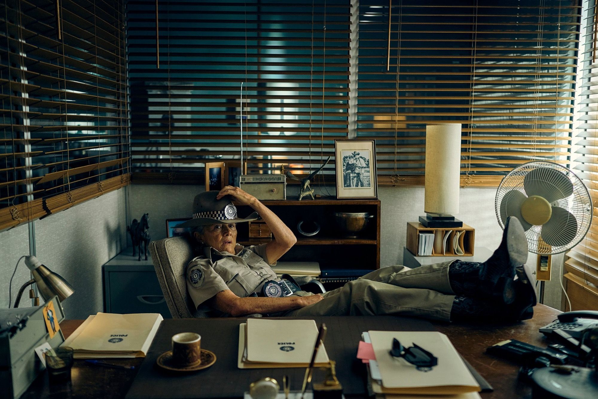 A police officer reclines at a cluttered desk, feet up, behind venetian blinds.