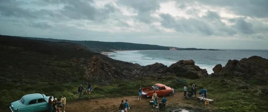 Young people and vintage cars gathered on a clifftop above the sea.