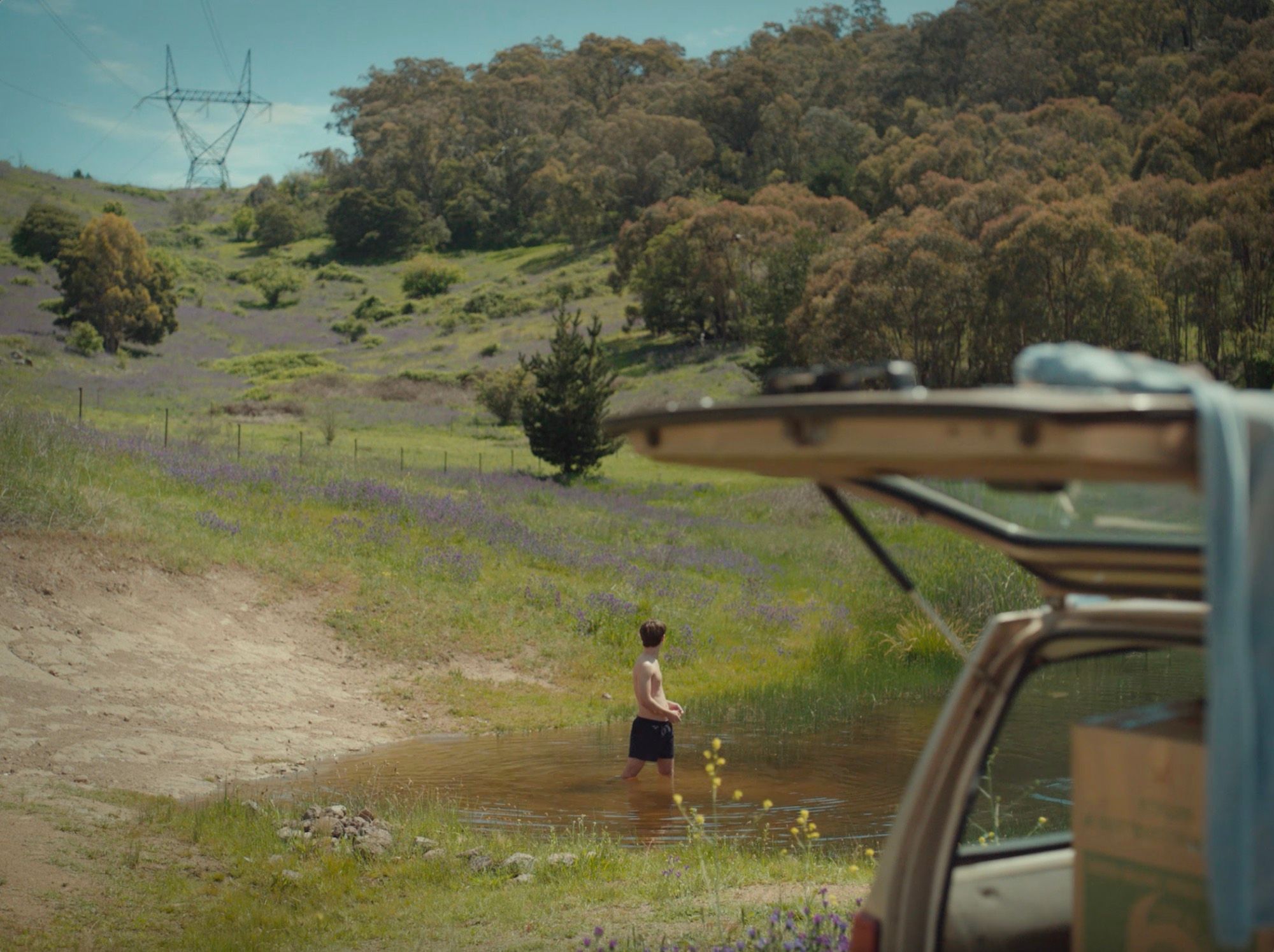 A boy wades into a dam below a hillside, beside an open car.